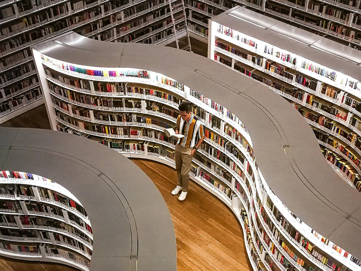 newcoUlibrary A man leans against an illuminated bookshelf, reading a book.