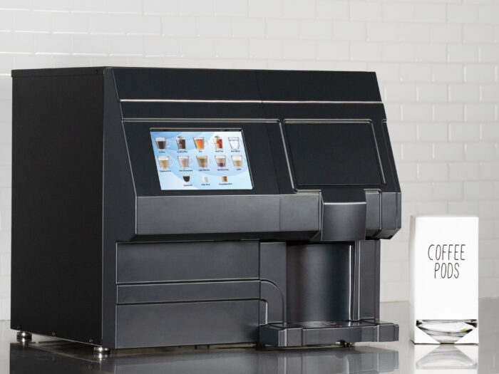 A black soft pod brewer, the CX-Touch Horizon, sits on a stainless counter in front of a white tile background. A box of coffee pods is visible.