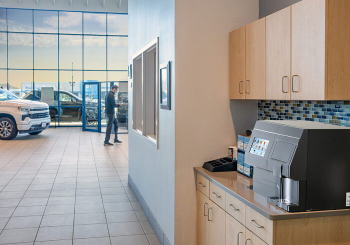 A black soft pod brewer sits on a break room counter in a car dealership as a man walks by in the background.